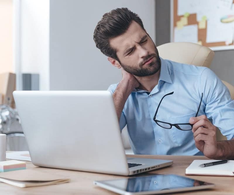 Hombre que sufre dolor de cuello mientras está sentado frente a un escritorio trabajando con una computadora portátil