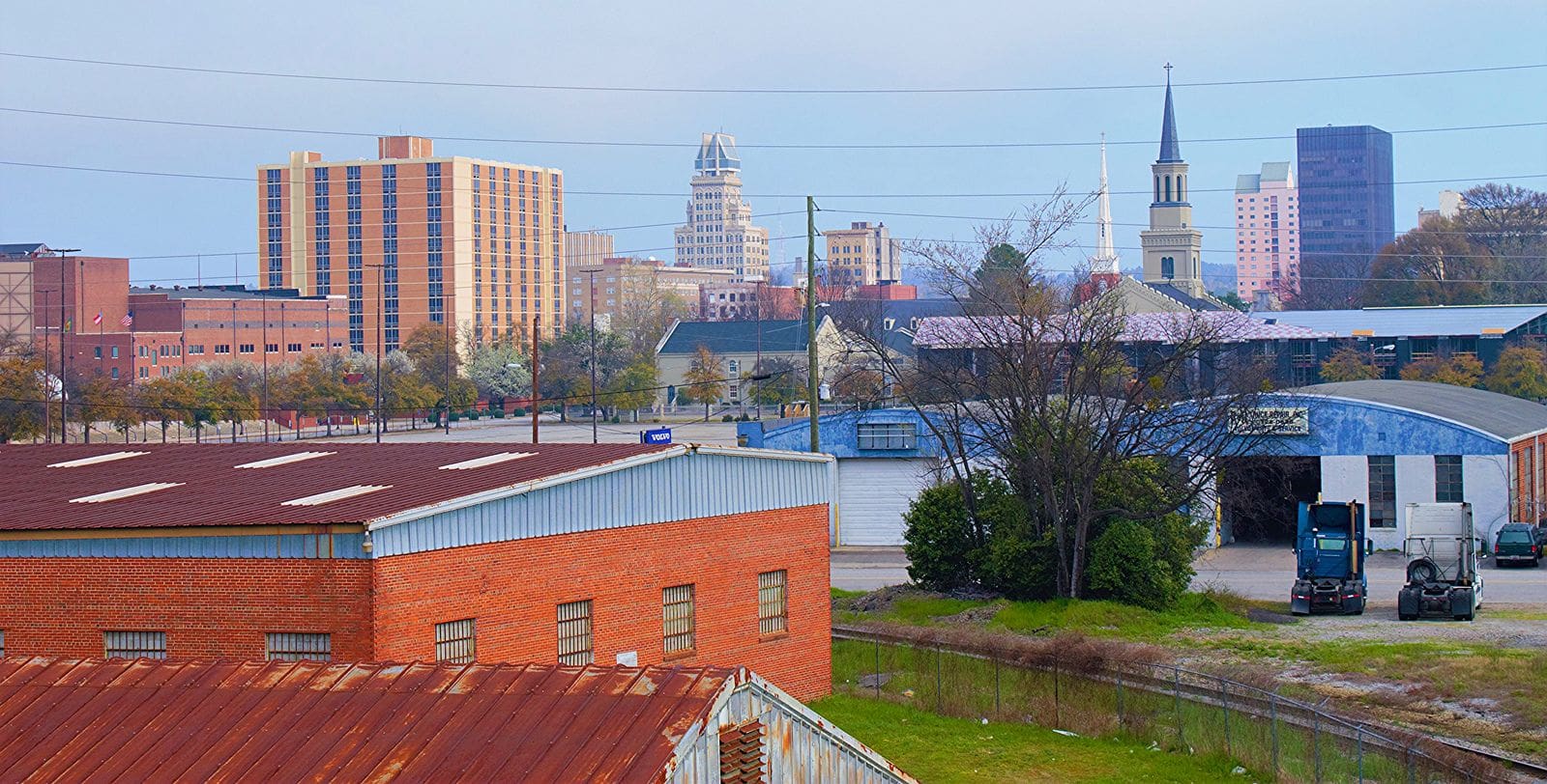 Augusta-area skyline with downtown buildings and a church steeple, viewed over industrial rooftops and rail-side warehouses.