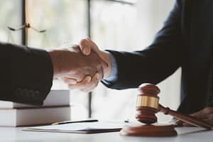 Two people in business attire shaking hands over a desk with a wooden gavel and documents in the foreground.