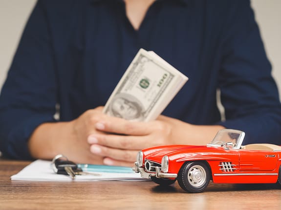 Mujer sosteniendo billetes de 100 dólares mientras está sentada en un escritorio con un Corvette rojo de juguete