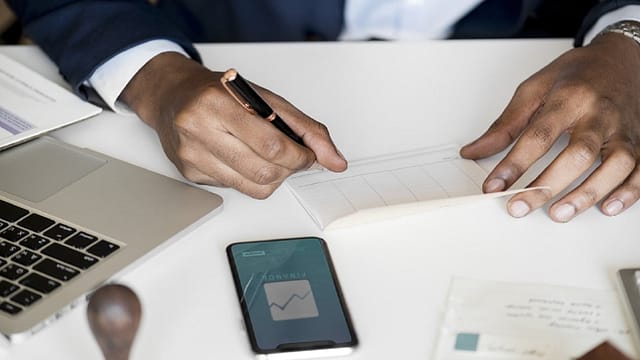 Person in a suit writing in a notebook on a desk with a laptop and a smartphone nearby.