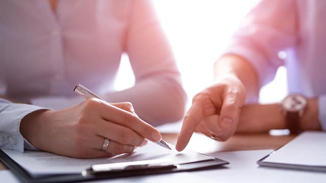 Close-up of two people reviewing documents at a desk, with one person writing and another pointing to the page.