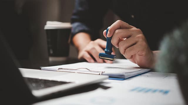 Close-up of a person pressing a rubber stamp onto documents on a clipboard, with a laptop and coffee cup visible in the background.