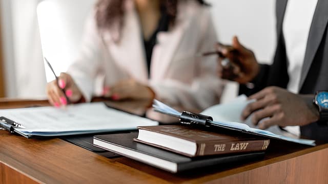 Two people reviewing and signing documents at a desk with a law book and clipboard in the foreground.
