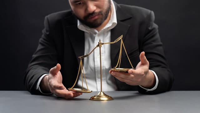 A bearded man in a suit holding out both hands on either side of a small brass scale of justice on a table.