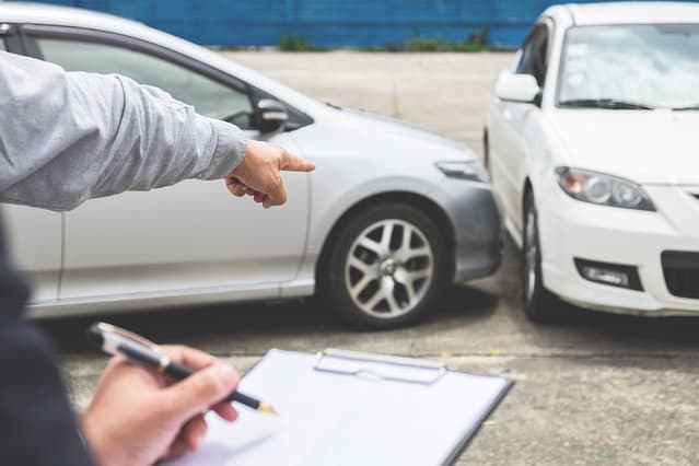 Person holding a clipboard and pen pointing toward two cars involved in a collision in a parking lot.