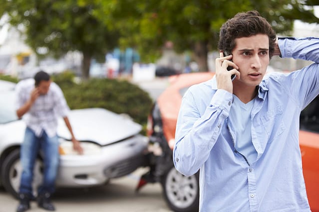 Young man talking on his phone with a worried expression at the scene of a two-car collision, with another person inspecting the damaged vehicles in the background.