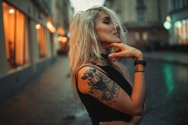 Young woman portrait with tattoo on shoulder standing on city street in evening. In background there are city lights. Laws in Texas for tattoos