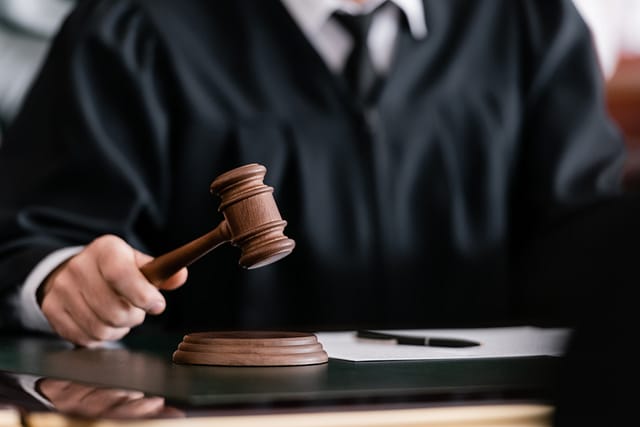Close-up of a judge in a black robe holding a wooden gavel above its sound block on a desk.