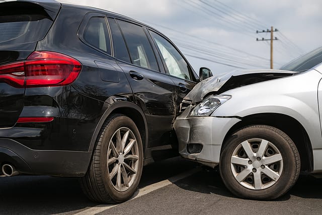 Silver car with front-end damage after a rear-end collision with a black SUV in a parking area.