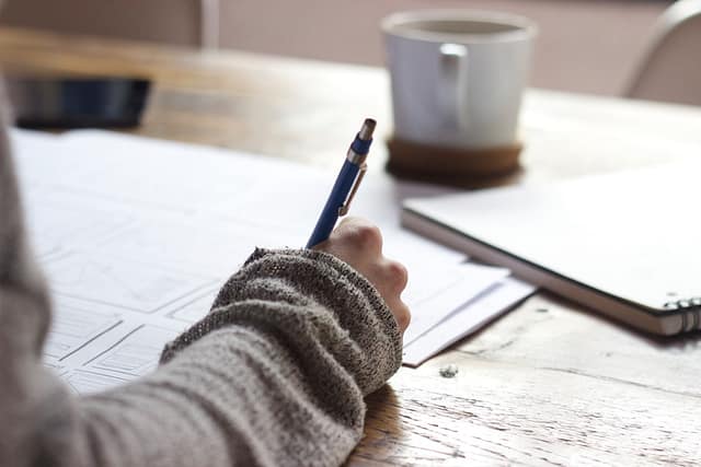 How Old Do You Have to Be to Get a Tattoo in Texas? Age, Consent & Legal Rules 3 Person in a knit sweater writing with a pen on paper at a wooden desk, next to a coffee mug, notebook, and phone.