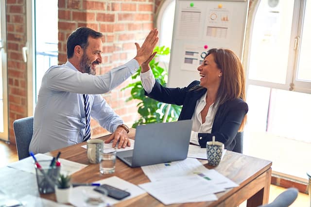 Two professionals giving a high five at a desk with a laptop and documents in an office setting.