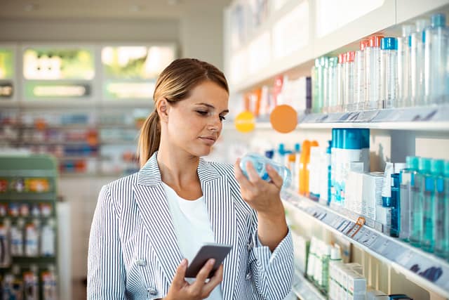 Woman reading a product label in a store aisle while holding a smartphone.
