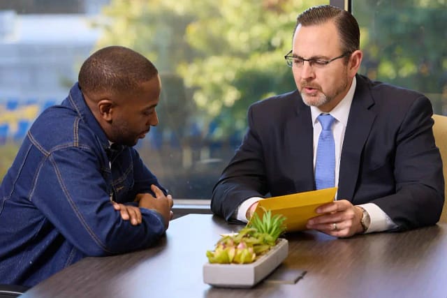 Who Is Liable for a Broken Sidewalk in Arizona? 4 Two men seated at a table, one in a suit holding a document and speaking, the other listening with hands folded.