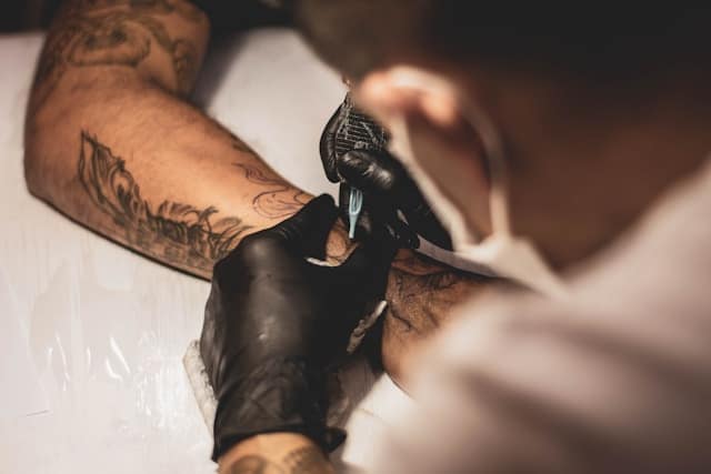 How Old Do You Have to Be to Get a Tattoo in Texas? Age, Consent & Legal Rules 4 Tattoo artist wearing a black glove using a tattoo machine on a client's tattooed forearm.