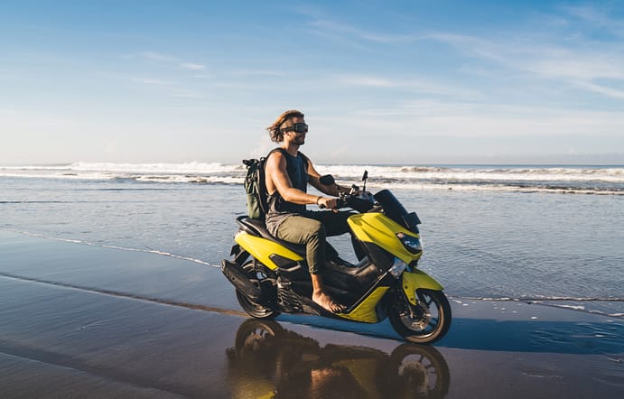 Longitud total de un viajero masculino con gafas montando una motocicleta sobre arena mojada cerca del mar agitado mientras mira hacia otro lado y descansa durante las vacaciones.