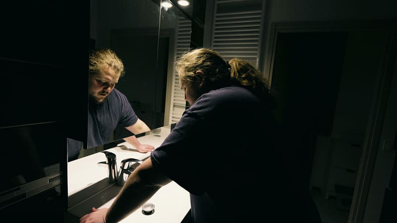 Hombre con angustia emocional apoyado en el espejo del baño que sufre una enfermedad mental solo en casa. Persona que siente soledad y depresión. Costo emocional de lesiones personales